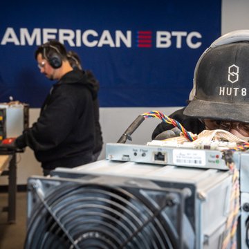 Two workers wearing 'HUT 8' branded caps and headphones work on what appears to be cryptocurrency mining hardware, with an 'AMERICAN BTC' banner visible in the background.