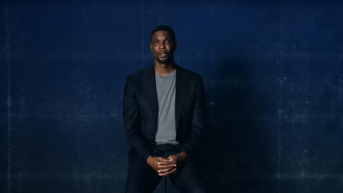 The image features a man wearing a suit and tie standing against a blue background with a white wall behind him. The man appears to be posing for a photo, possibly as part of an advertisement or promotional campaign related to Bitcoin.