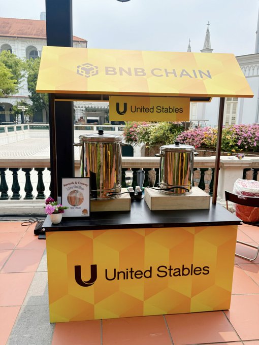 A yellow promotional booth branded with "BNB CHAIN" at the top and "U United Stables" on the front and a side sign, featuring two large silver beverage dispensers and a smaller sign for "Smooth & Creamy Corn Stollen."