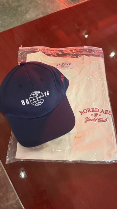 A navy blue "BAYC" cap rests atop a folded "Bored Ape Yacht Club" t-shirt sealed in plastic packaging, displayed on a glossy red surface—physical merchandise embodying the post's claim that IRL presence is the ultimate flex for Bored Ape Yacht Club members.