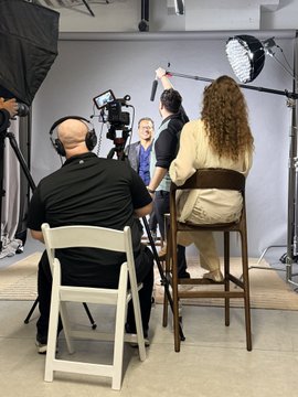 A behind-the-scenes view of a film or photo set with crew members, cameras, lighting equipment, and two seated subjects facing each other.