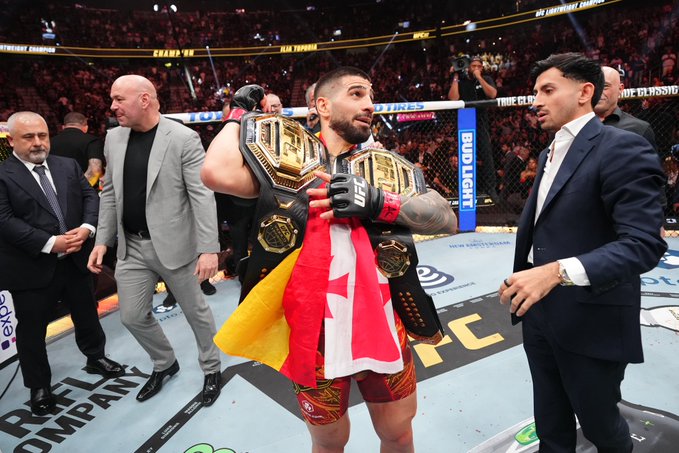 The image shows a man holding two championship belts, one red with a yellow star and the other black with a gold star. He appears to be celebrating his victory at an event or competition. The surrounding people are dressed in suits and ties, indicating that this is likely a formal occasion. The post about the UFC Freedom fights presents the man's achievement as a fighter who has won multiple titles within the organization.