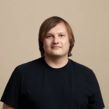 A portrait photograph of a man with shoulder-length light brown hair wearing a plain black t-shirt against a beige background.