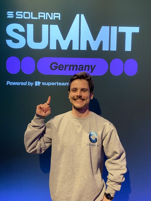 The image features a man standing in front of a wall with a sign that says "Solana Summit Germany" written on it, indicating an event or gathering related to the Solana Summit. The man appears to be posing for a photo, possibly at a convention or exhibition. He is wearing a grey sweatshirt and has his finger extended upwards, as if he's giving an order or making a gesture of approval. In the background, there are other people present who might also be attending the event.
