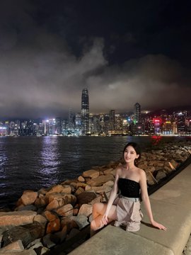 A young woman in a strapless top and light skirt poses along a rocky waterfront at night, with a stunning illuminated city skyline reflected across the harbor behind her. The scene of a leisurely evening stroll complements the post's casual mention of exploring newly opened crypto communities like OK Star and Binance Square.