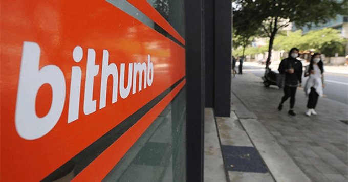 Photograph of a Bithumb cryptocurrency exchange storefront sign with two pedestrians walking on the sidewalk beside it.