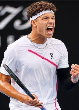A young tennis player with curly hair, wearing a white headband and white "On" branded shirt, shouts passionately while gripping a racket and clenching his fist in a victorious celebration. The image relates to the post about the large $92,000 bet placed on Ben Shelton to win the US Open against Alexander Zverev, capturing the intensity and competitive spirit associated with such high-stakes wagers.