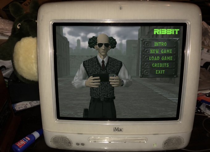 A vintage white iMac CRT monitor displays the main menu of a video game called "RIBBIT," featuring a bald character in sunglasses and a sweater vest holding a device, with menu options for INTRO, NEW GAME, LOAD GAME, CREDITS, and EXIT visible on the right side of the screen.