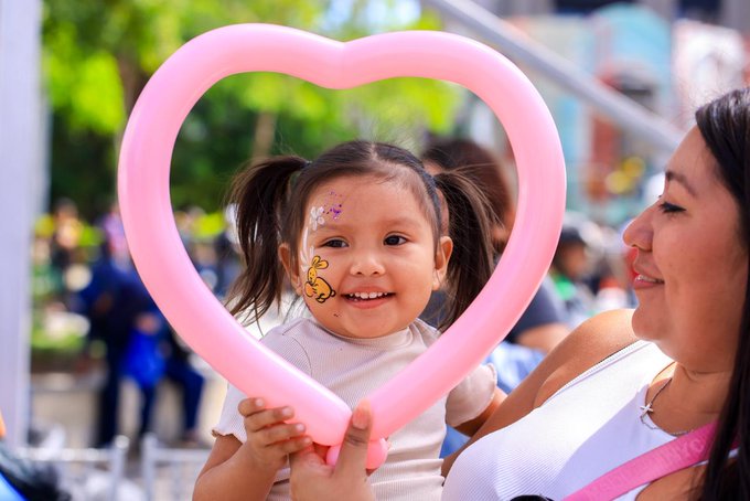 A young girl with pigtails and a Winnie the Pooh face sticker smiles brightly while holding a pink heart-shaped balloon, as a woman beside her gazes at her warmly. The image's joyful, optimistic tone aligns with the post's celebration of El Salvador's new era of relentless optimism, visually echoing the message that joy and positivity are by design.

No visible text, numbers, tickers, or names appear on the balloon or clothing in the image.