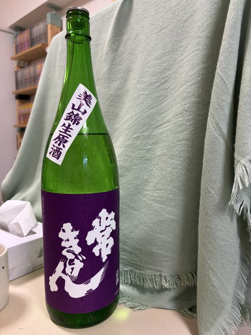A green glass bottle of 常きげん (Jou Kigen) sake with purple and white labels stands on a table against a draped green fabric backdrop, the large 1.8L isshobin size confirming the poster's note about buying the larger bottle for better value.