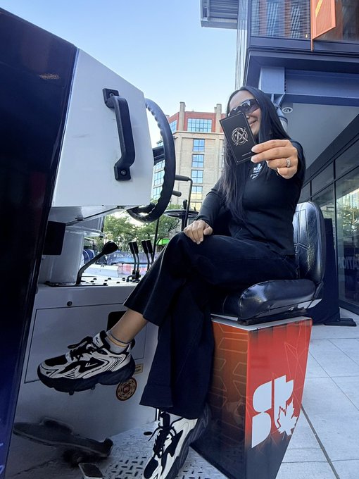 A woman in black attire and sunglasses sitting on a Dunkin' Donuts-branded seat while taking a mirror selfie outside a brick building.