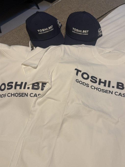 Two white t-shirts and two dark blue caps are laid out, all featuring the text 'TOSHI.BET' and 'GODS CHOSEN CASINO'.