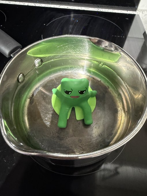 A small green clay figurine with an angry face sits on a green disc inside a metal saucepan on a stovetop.