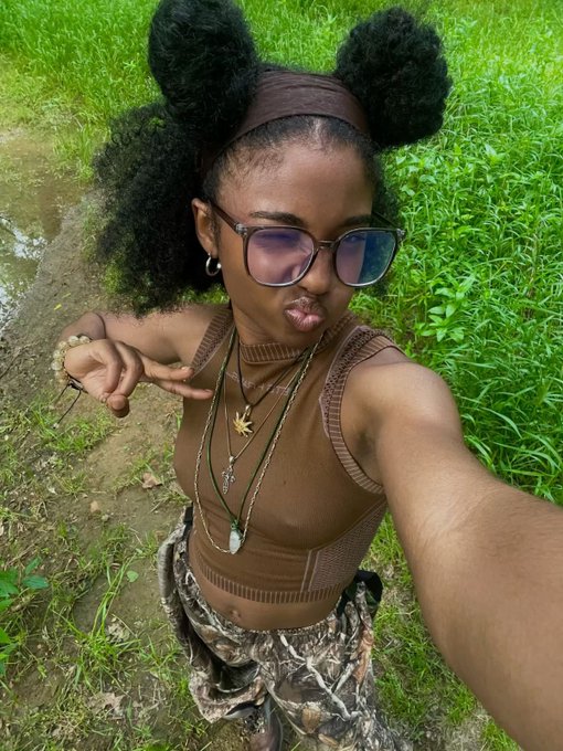 The image features a young woman with long hair wearing glasses and a brown shirt. She has her hair styled into two pigtails and is posing for a photo, holding up one finger as if she's giving a thumbs-up or making a peace sign. The background of the image shows grassy areas, suggesting that they are in an outdoor setting like a park or field. There is also text present in the image, which reads "happy weekend umu Chineke touch grass, explore $RIVER mind your business and make monies".