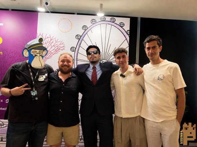 A group of five people pose together at what appears to be a crypto/blockchain event in Singapore, with one man in a suit wearing a lanyard standing among casually dressed attendees. The backdrop features cartoon graphics including a character resembling a captain and a ship wheel, and the post suggests this is proof of attendance at a Solana-related conference, with a "BRICS" tag visible in the corner.