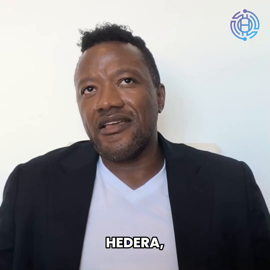 The image features a man wearing a black suit jacket, sitting down against a white background. The man has short hair and appears to be looking off into the distance or at someone else who is not visible in the frame. There are no texts or other objects present in the image.
