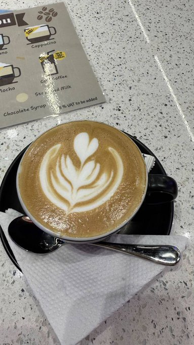 A beautifully crafted cappuccino with intricate latte art sits on a saucer beside a coffee menu referencing "Cappuccino," "Soft Coffee," "Sterilized Milk," and "Chocolate Syrup" — paired with a caption about starting the week fresh, using the morning coffee ritual as a metaphor for renewed energy and long-term interest in River (RiverdotInc).