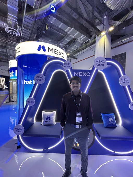 A smiling man in a maroon shirt standing in front of an illuminated blue MEXC booth at a crypto conference, featuring sections for MEXC Card, Stocks, and Earn.