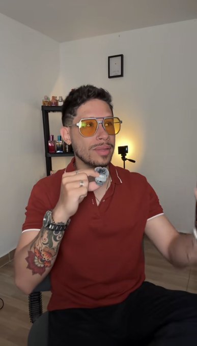 A man wearing orange-tinted sunglasses and a red polo shirt, with a tattoo on his arm, sitting indoors and holding a small object near his face.