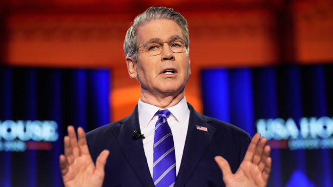 Treasury Secretary Scott Bessent speaks at a podium with a "USA IHO" backdrop visible behind him, gesturing with both hands raised. The image accompanies discussion about his anticipated announcement of expanded Treasury buybacks aimed at managing long-term yields and improving liquidity conditions for risk assets like Bitcoin.
