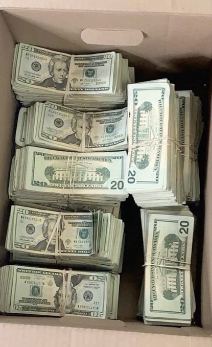 An open cardboard box filled with multiple banded stacks of US twenty-dollar bills, showing the obverse (Andrew Jackson) and reverse sides of the currency.
