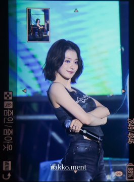 A concert photo of a female K-pop idol with short dark hair wearing a black sleeveless outfit, posing with crossed arms and holding a microphone, with 'makko.ment' watermark text visible.