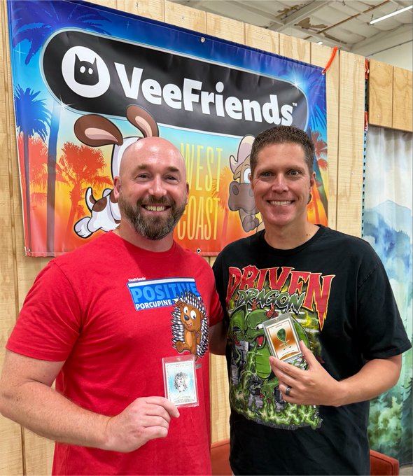 Two smiling men pose in front of a "VeeFriends" banner while proudly displaying collectible cards—the man on the left in a red "Positive Porcupine" shirt holds a VeeFriends card, and the man on the right in a "Driven Dragon" shirt holds a Series 1 Spec card, marking a meaningful NFT-to-collectible milestone celebrated between friends in the #VFam community.