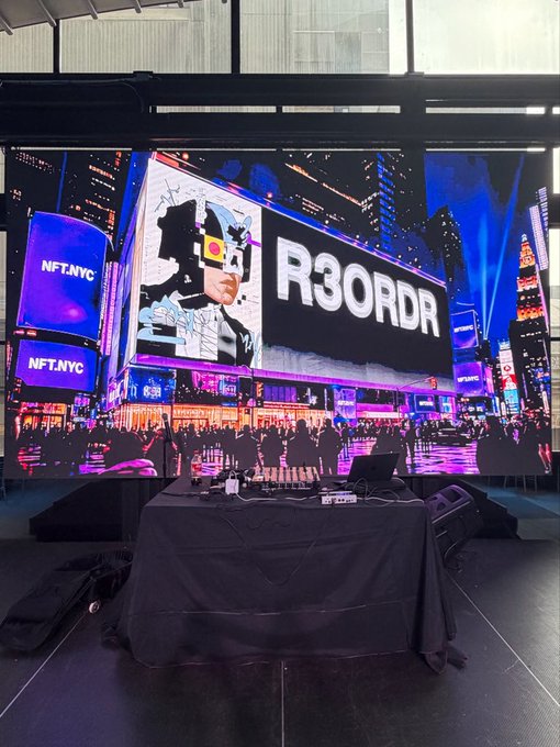 A DJ setup with turntables in front of a large screen displaying 'R3ORDR' alongside NFT.NYC branding and a stylized Times Square background.