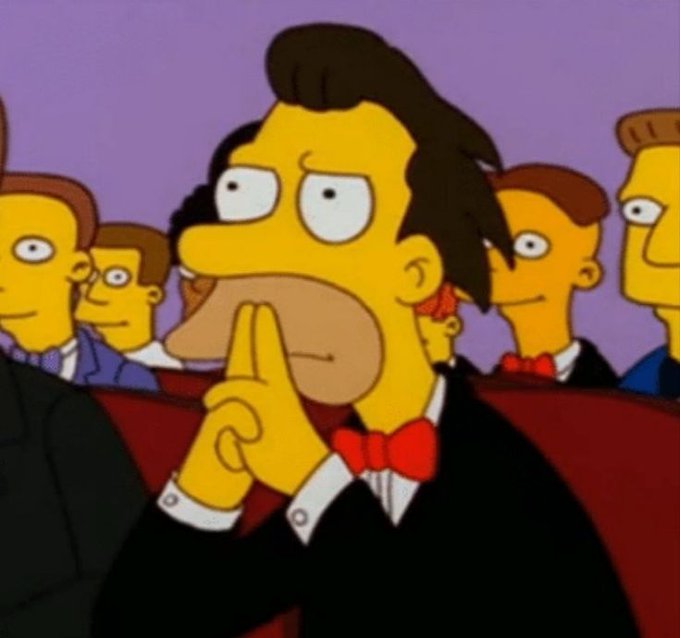 A character from The Simpsons, dressed in a black tuxedo with a red bow tie, sits in red theater seats with a thoughtful expression, finger on chin, surrounded by other formally dressed characters.