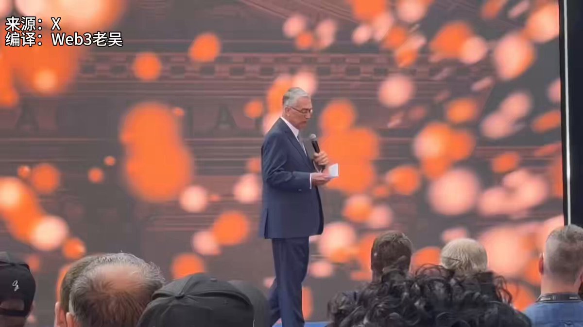 The image shows a man standing at a podium, giving a speech or presentation to an audience. The speaker appears to be wearing a suit and tie, suggesting a formal event or occasion. The audience consists of several people gathered around the stage, attentively listening to the speaker's words.