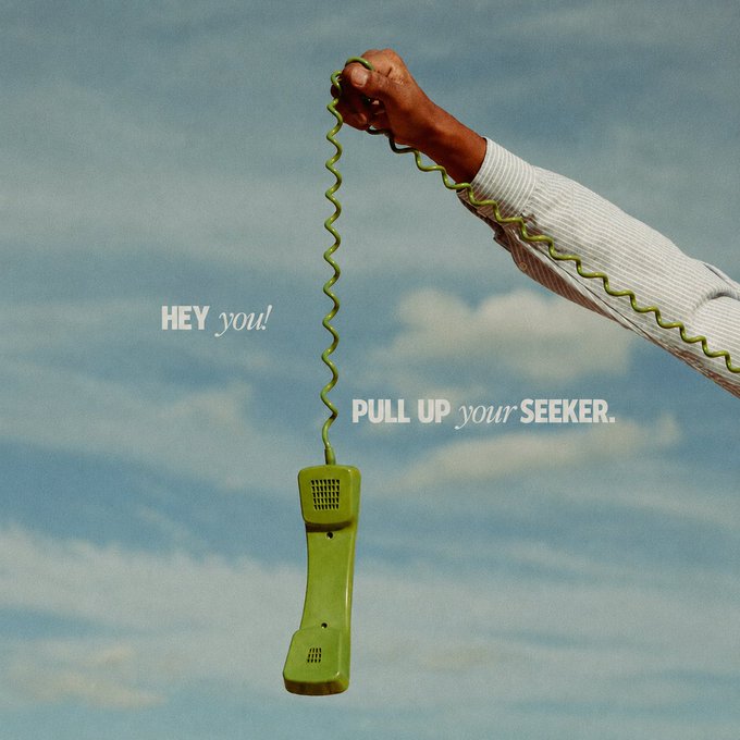 A hand holds the green coiled cord of a wall-mounted telephone receiver against a blue cloudy sky, accompanied by the text 'HEY you! PULL UP your SEEKER.'