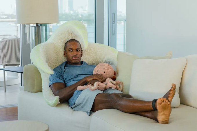 The image shows a man sitting on a couch with an octopus plush toy between him and another person who is not visible in the frame. The scene appears to be set in a living room, where there are two chairs nearby. A lamp can also be seen in the background. The image seems to be related to a post about a man dumping his bag on social media or an advertisement for a product named "if you're selling your $HYPE just because this guy dumped his bag".