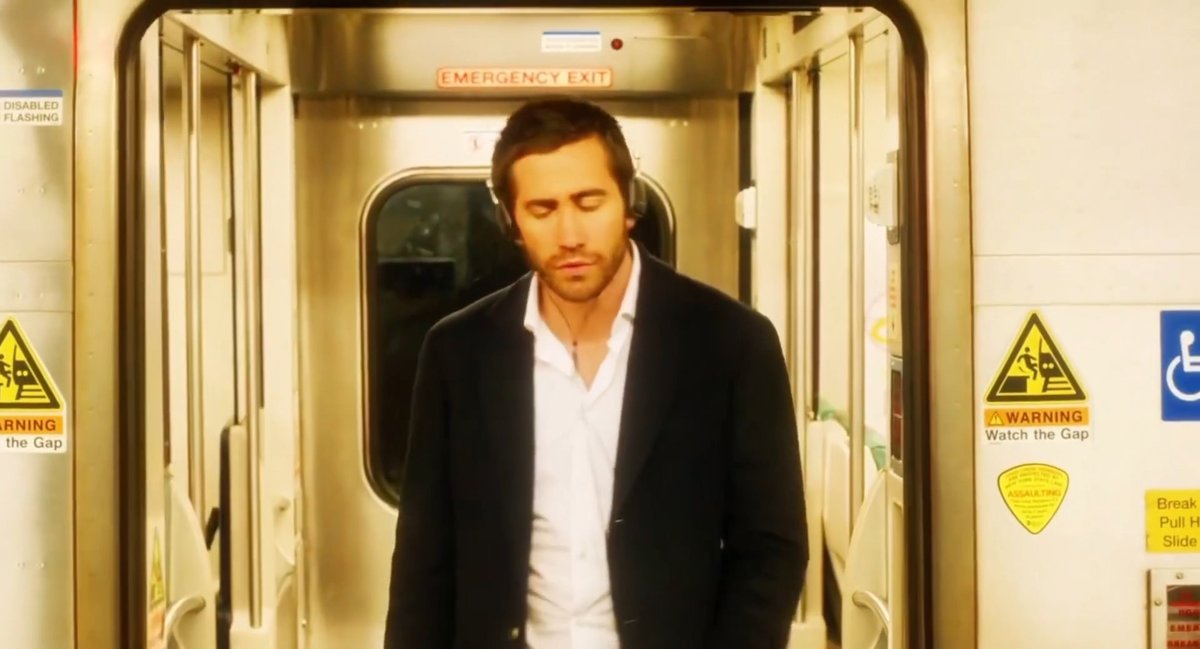 A man in a dark suit and white shirt wearing headphones, standing with his eyes closed in a subway car near an 'EMERGENCY EXIT' door with 'Watch the Gap' warning signs visible.