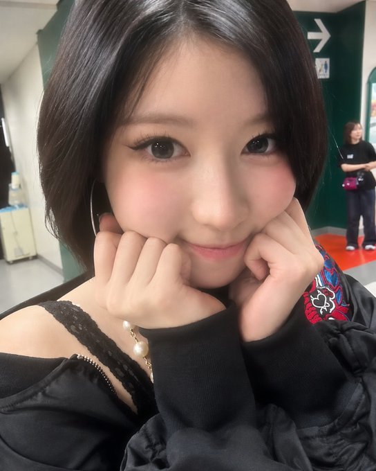 A young woman with dark hair poses cutely, resting her chin on her hands with rosy cheeks and a sweet smile, wearing a black top with a pearl necklace. In the background, a green wall displays a white directional arrow, while another person stands in a hallway. The "#ASA" hashtag suggests this is a post honoring or featuring the South Korean singer Asa (also known as Yeri), known by fans as a charming "pretty bunny."