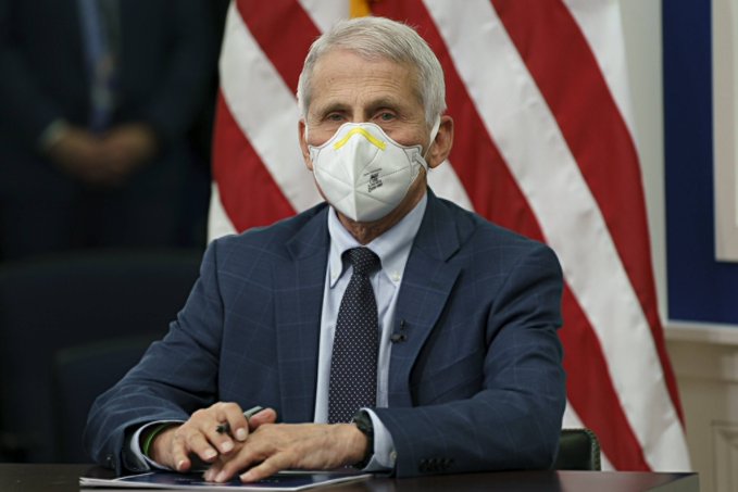 Dr. Anthony Fauci, wearing an N95 mask and dark blue suit, sits at a desk with an American flag behind him, in what appears to be an official government setting.