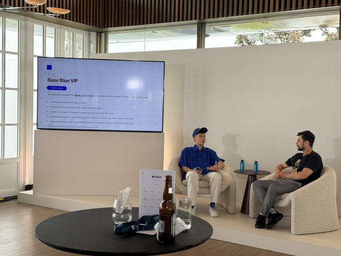 Two men seated in chairs on a stage having a discussion, with a presentation screen behind them displaying 'Base Blue VIP' program details for Base's ecosystem.