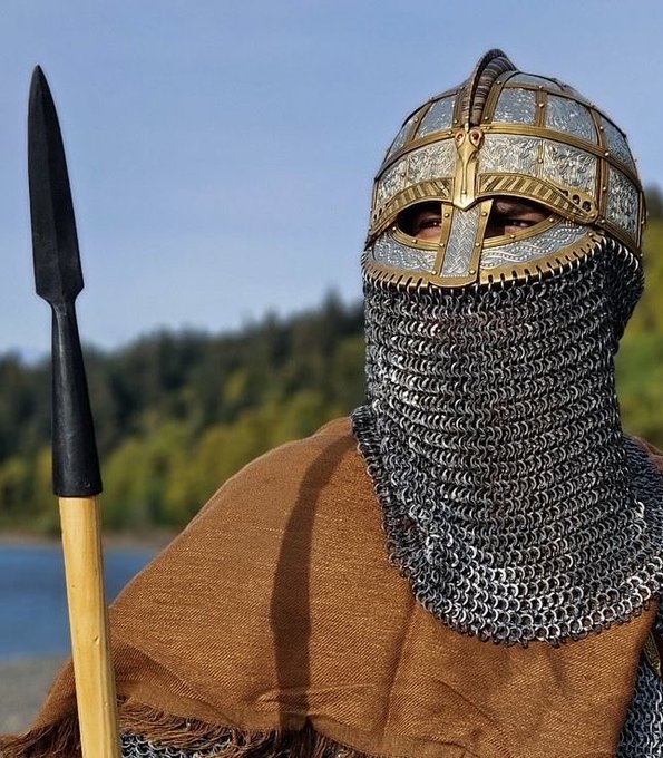 A person dressed in medieval Viking attire, wearing a chainmail coif beneath an ornate steel helmet with brass trim, holds a wooden-handled spear against a backdrop of a blue sky and forested lakeshore.