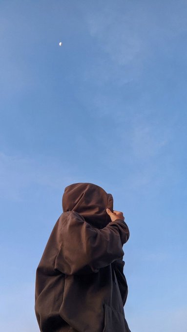 A person wearing a brown hooded robe, photographed from below, pointing up at a small crescent moon in a clear blue sky.