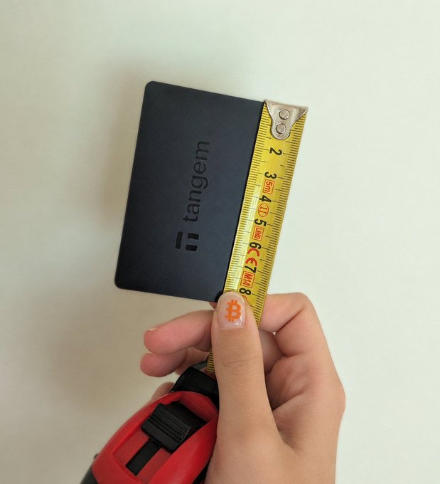 A hand holds a Tangem hardware wallet against a yellow tape measure, showing it's approximately 8 cm tall—matching the claim that "it only takes 8cm to make a woman happy," humorously highlighting the compact size of the crypto wallet.
