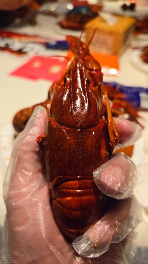 A close-up of a hand wearing a clear plastic glove holding a freshly cooked, bright red cooked crawfish or crayfish, with more lobed shells on the table in the background.