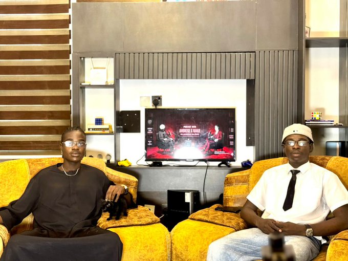 Two men sit in yellow chairs in a living room watching a screen that displays what appears to be a Red Bull racing event.