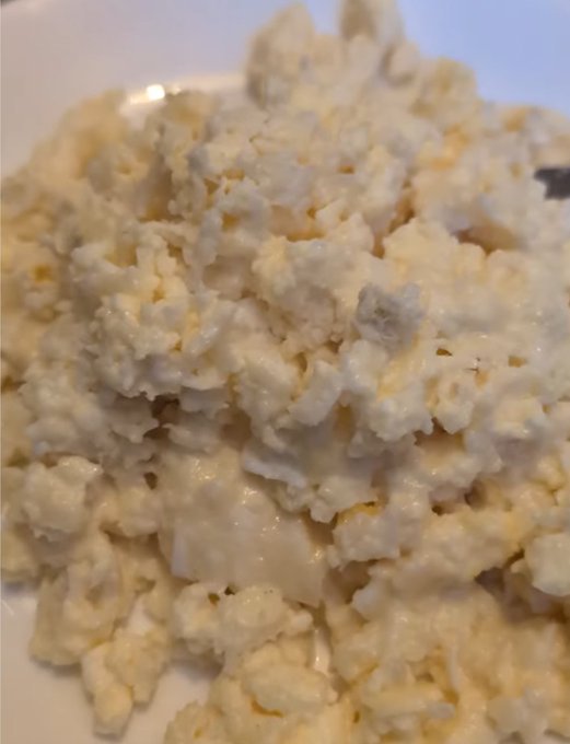 A close-up photograph of a serving of crumbly, pale yellow and white food, possibly cottage cheese or scrambled eggs with cheese.