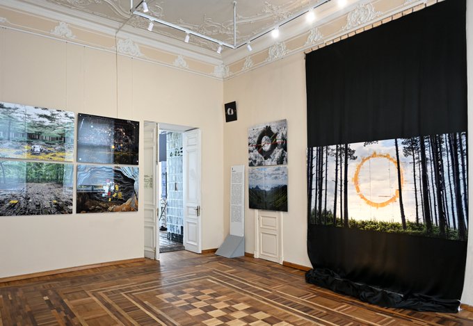 An art gallery interior with parquet flooring and ornate ceiling moldings, displaying large photographic prints on the walls including one showing a circular orange shape over a forest.