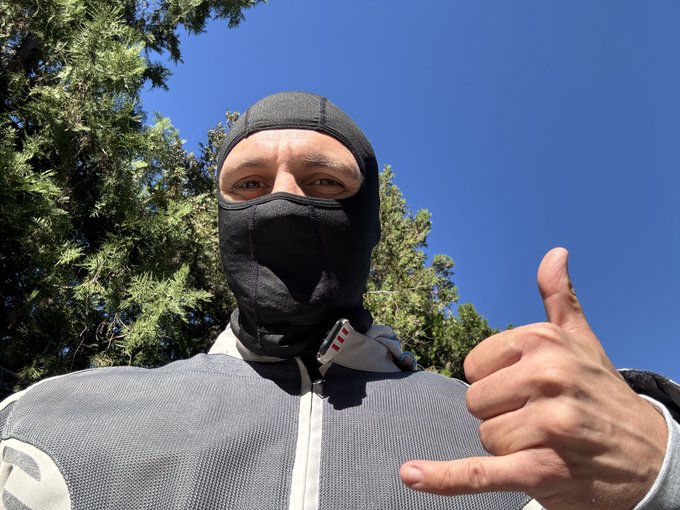 A man wearing a black balaclava and a gray jacket gives a shaka hand sign while standing outdoors under a clear blue sky with green trees in the background.