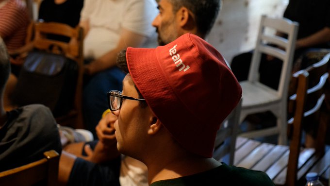 A side view of a man wearing glasses and a red beanie with the word 'LUCAS' in white letters, sitting among other people in what appears to be a social gathering.