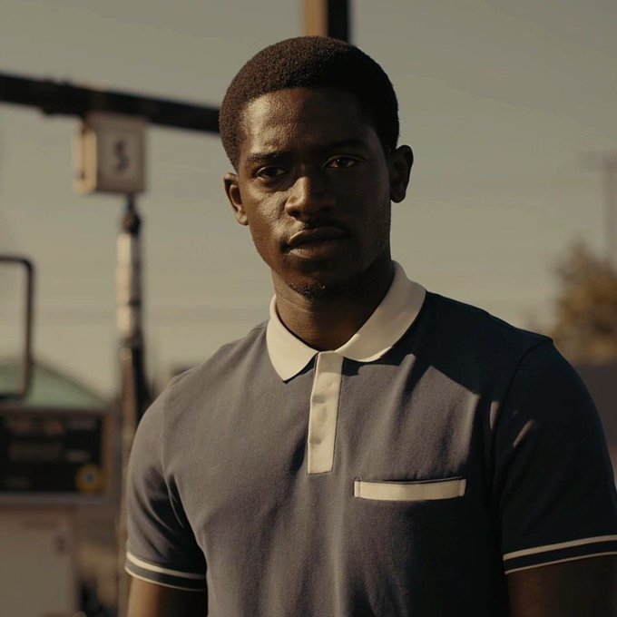 A portrait of a young Black man wearing a dark polo shirt with white trim, standing outdoors near a gas station pump during the golden hour.