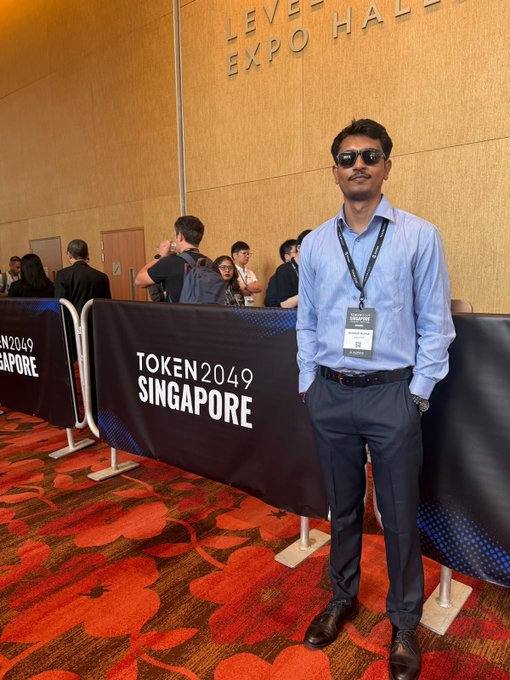 A man wearing sunglasses and a blue shirt with a lanyard stands in front of a 'TOKEN2049 SINGAPORE' banner at the Level 4 Expo Hall.