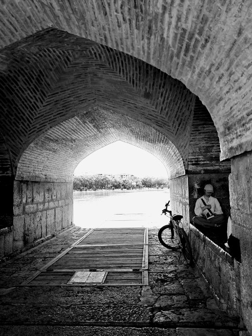 A black and white photograph taken from inside a brick archway looking out toward a body of water, showing a person in a hard hat sitting on the right with a bicycle nearby.