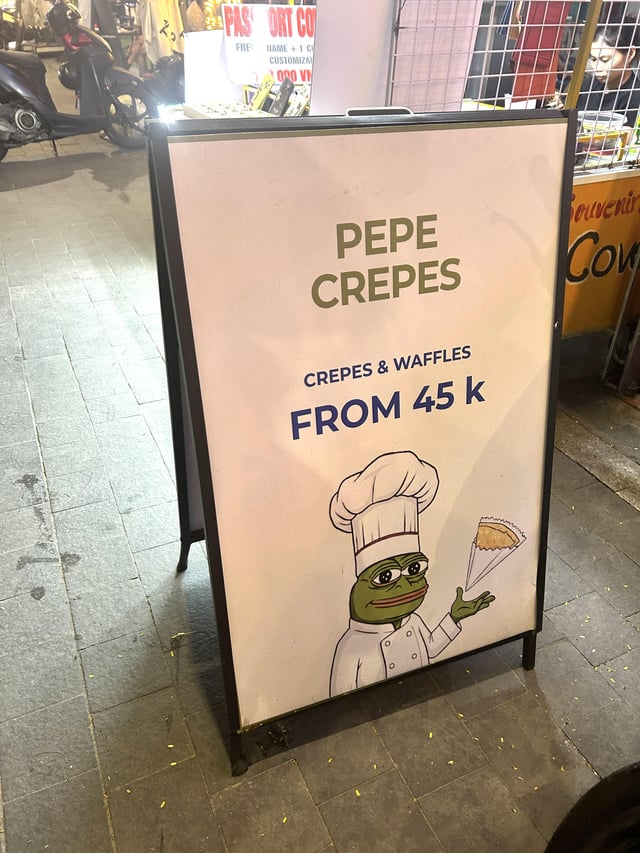 An A-frame sidewalk sign for "PEPE CREPES" advertises crepes and waffles starting from 45k, featuring a Pepé the Frog mascot dressed as a chef holding a crepe—fittingly playing on the $PEPE meme cryptocurrency for a real-world themed food stand.