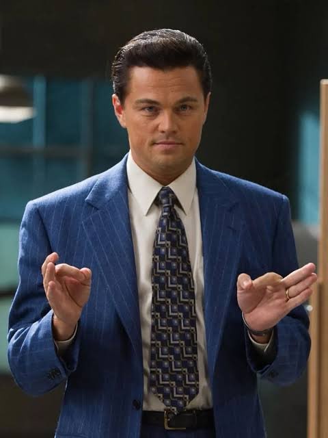 Leonardo DiCaprio stands in a blue pinstripe suit with a patterned tie, holding both hands up with fingers pinched together in the "Money Money" gesture from The Wolf of Wall Street.